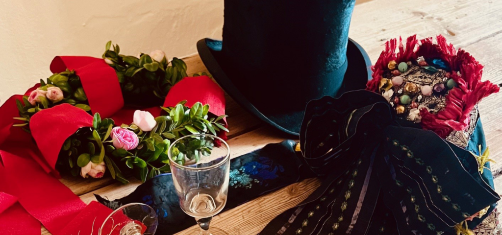 Stilleben med vinglas, buksbomkrans, høj sort silkehat og kyse på en bordplade af råt træ