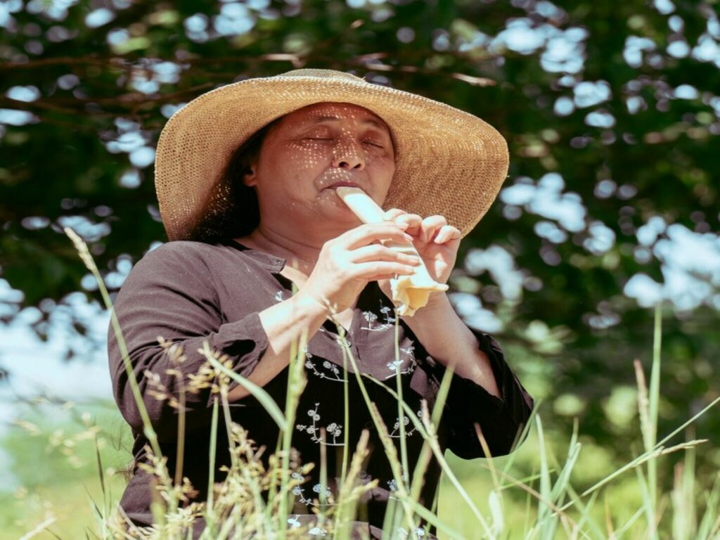 Kvinde med stråhat spiller på fløjtelignende genstand midt i naturen.